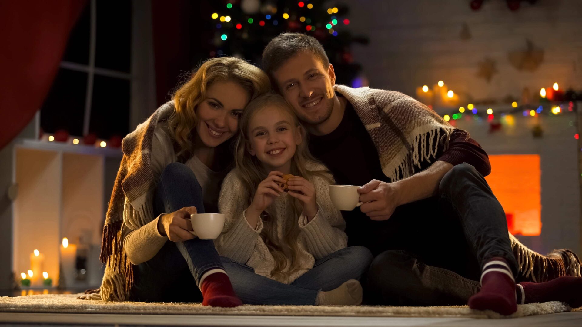 Eine Famlie sitzt angekuschelt auf dem Boden und im Hintergrund sieht man Kerzen und Lichter.